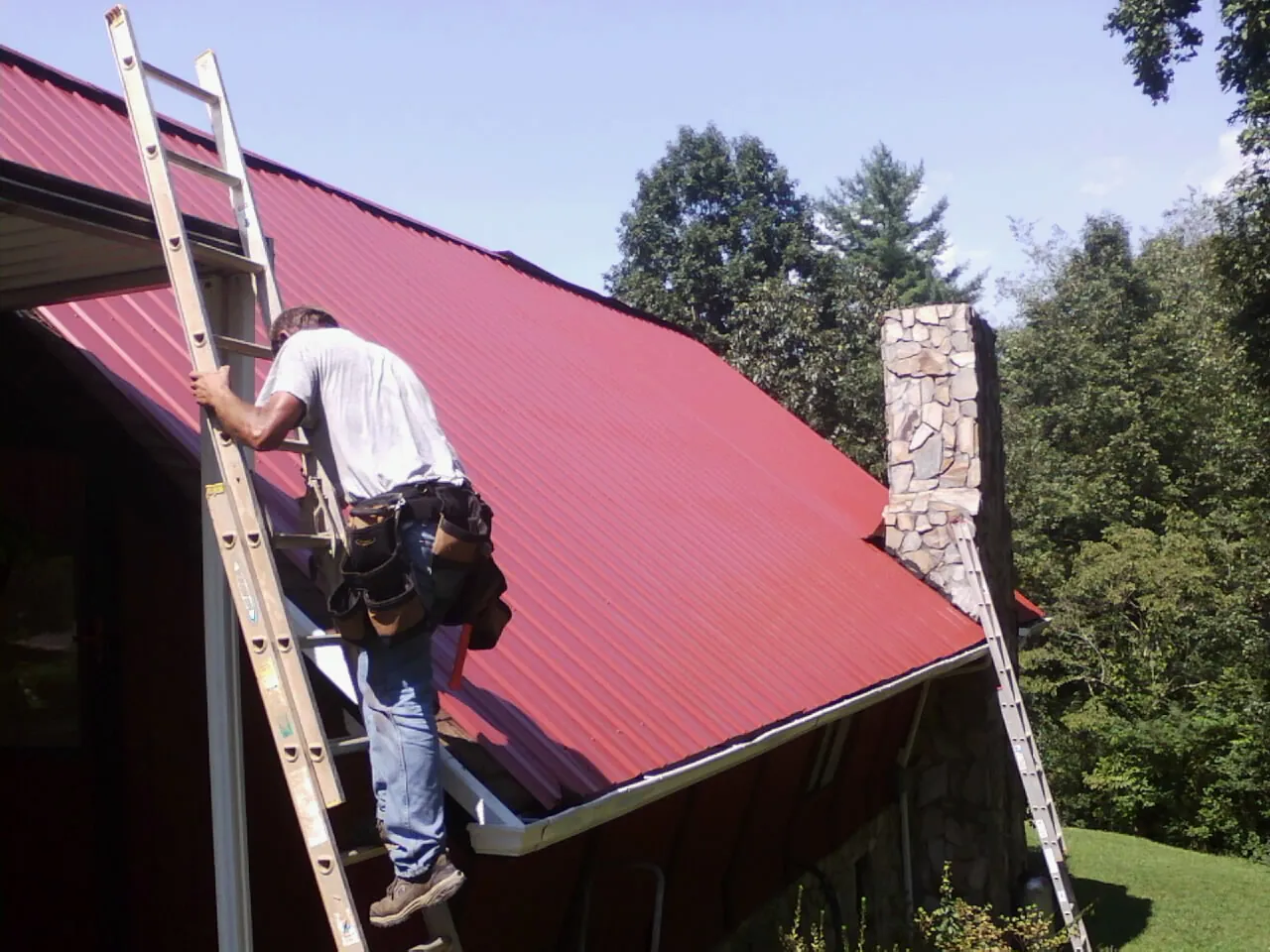 Roofer on a ladder beside a red metal roof and stone chimney — Soffit & Fascia Repair by Roofing Brothers in Saint Bonifacius